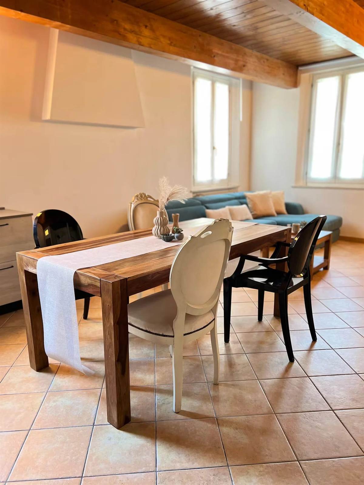 Zona pranzo e soggiorno di Domus Cerchio 19: tavolo in legno, sedie bianche e nere, divano azzurro, due finestre e travi a vista