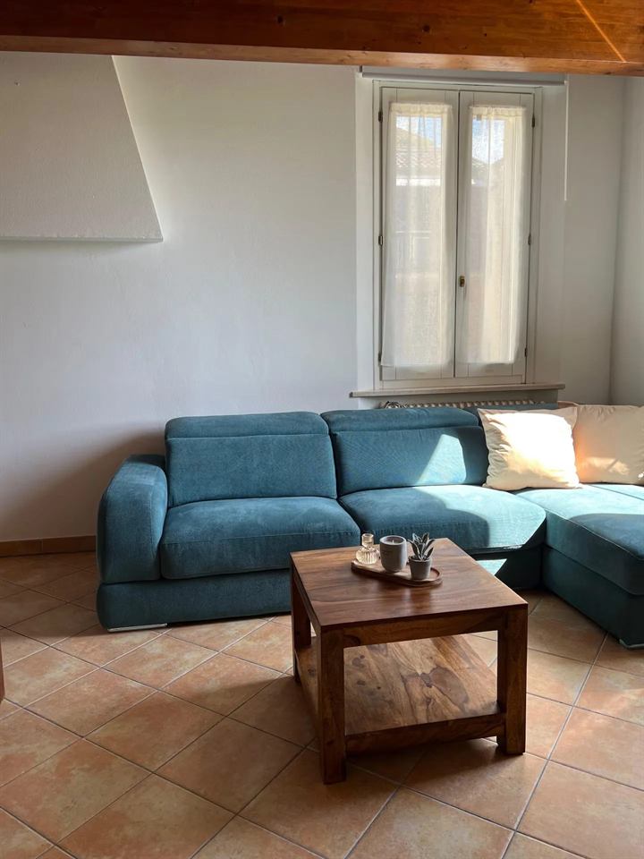 Divano angolare azzurro sotto la finestra con tende in lino di Domus Cerchio 19, tavolino in legno con candela, pavimento in cotto